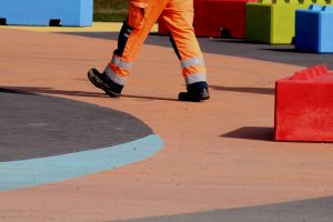 A peron walks through a concrete plaza wearing orange safety pants and black boots. In the background are red, green and blue life-sized building blocks.