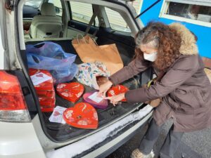 Image of a woman wearing a brown winter jacket and a mask leaning over the trunk of a silver unidentified SUV taping printed cards with hearts on white paper onto red, heart-shaped boxes of chocolates..