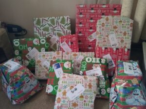 Image of presents wrapped in a multitude of colorful paper including white, red, and green. The presents are stacked leaning against a white wall on top of a beige carpet.