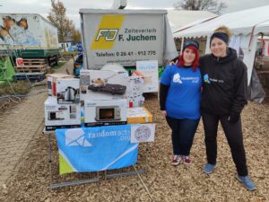 Two women stand arm in arm to the left of a table with many donated items in cardboard boxes. A Random Acts banner is displayed in front of the table.