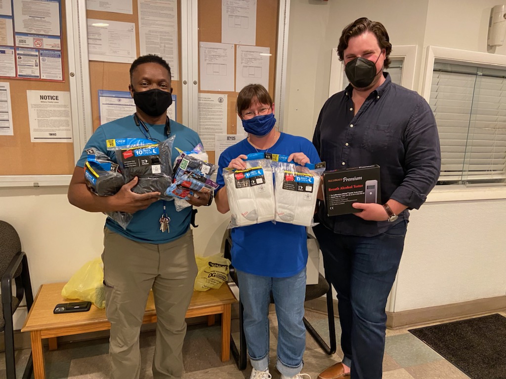 Three people are standing next to each other. On the left stands a man with a blue shirt and a black medical masks. He is standing left to a woman with a azure blue t-shirt. To her right stands another man with a dark longarm-shirt. All three are wearing black medical masks and hold donations (socks and underwear) in their hands