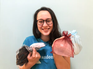 Image of Regional Lead Rowan Meredith smiling at the camera wearing her blue Random Acts t-shirt while holding chestwear such as breast forms, bras, and binders in sheer bags. She stands against a white background.