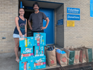 Two people stand in front of a building. Diaper boxes and shopping bags are in front of them.