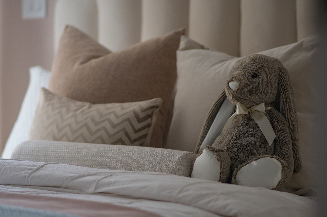 A toy bunny sitting on a freshly made bed with tan and grey pillows.