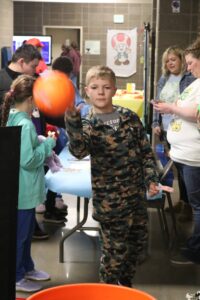 a boy in camo tossing a ball toward the basket at the front of the photo