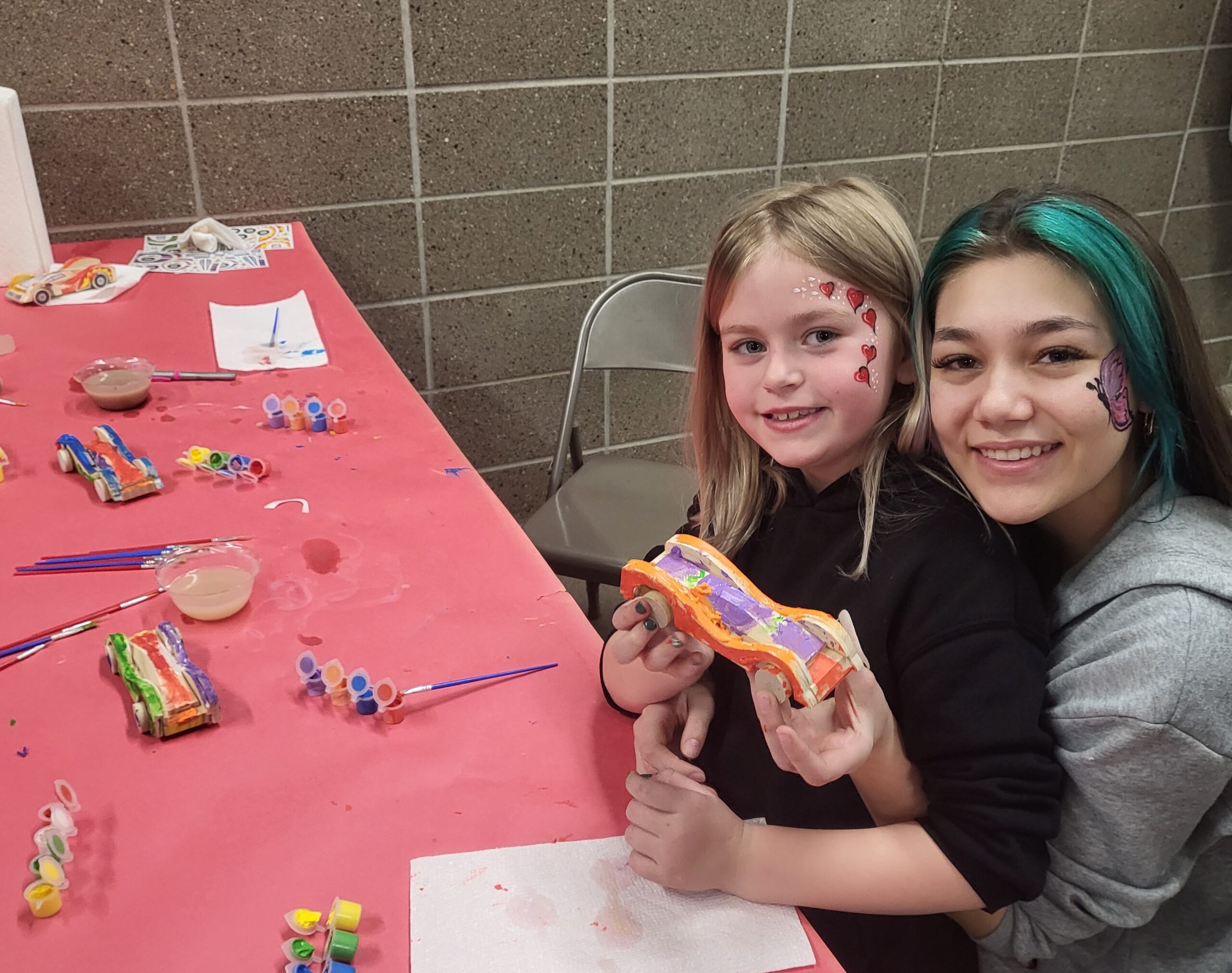 a young girl with blonde hair and pink face paint on her cheek holds her finished toy car while sitting in the lap of another girl with green and black hair, and a dolphin painted on her face