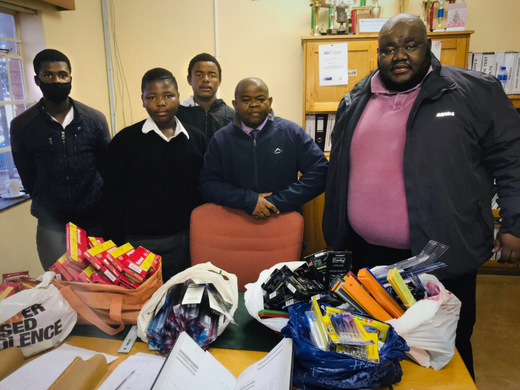 Five people stand behind a desk that has bags full of school supplies on it.