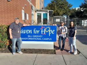 Three people stand around a big blue sign saying "Haven for Hope". To the right of the sign stand a man with a checked shirt, cap, and colourful lanyards. Next to him stands a young woman with brown hair and a black Random Acts T-Shirt. To the left stands a man in a striped Shirt and a lanyard.